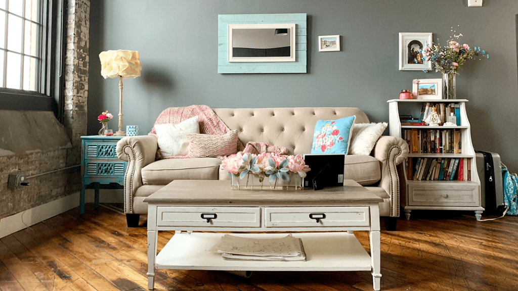 A livingroom with an off white sofa and pink and blue spring themed pillows, an off white coffee table with a pink flower arrangement